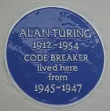 A Blue plaque on a white wall with the words "Alan Turing 1912–1954 CODE BREAKER lived here from 1945&nbsp;– 1947