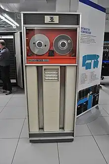 A large cabinet, about the size of an upright refrigerator, with a glass-covered top part holding two reels of magnetic tape, and a bottom part with control buttons framed by vertical channels.